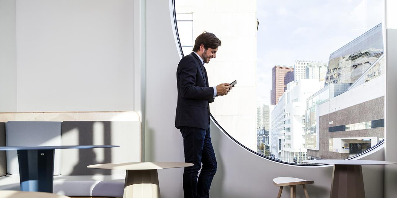 intérieur salon jeune homme business consulte tablette adossé grande vitre arrondie paysage urbain